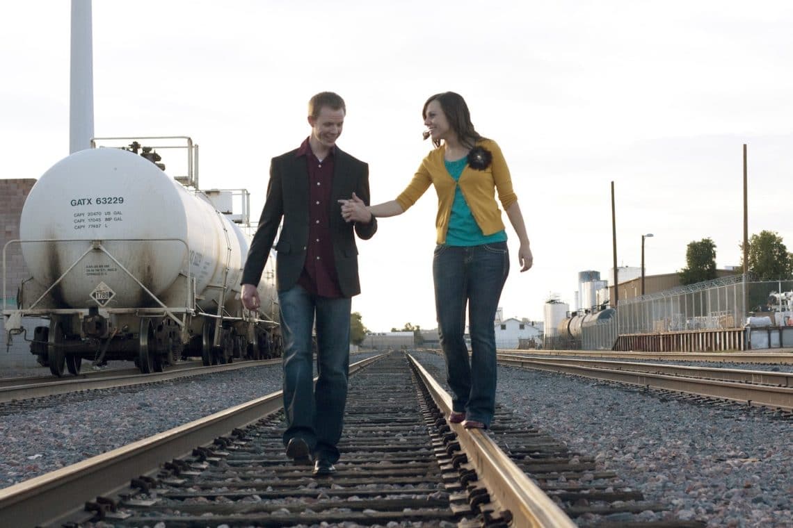 Couple pictures at a train station with the husband holding the wife\'s hand on the train tracks. 