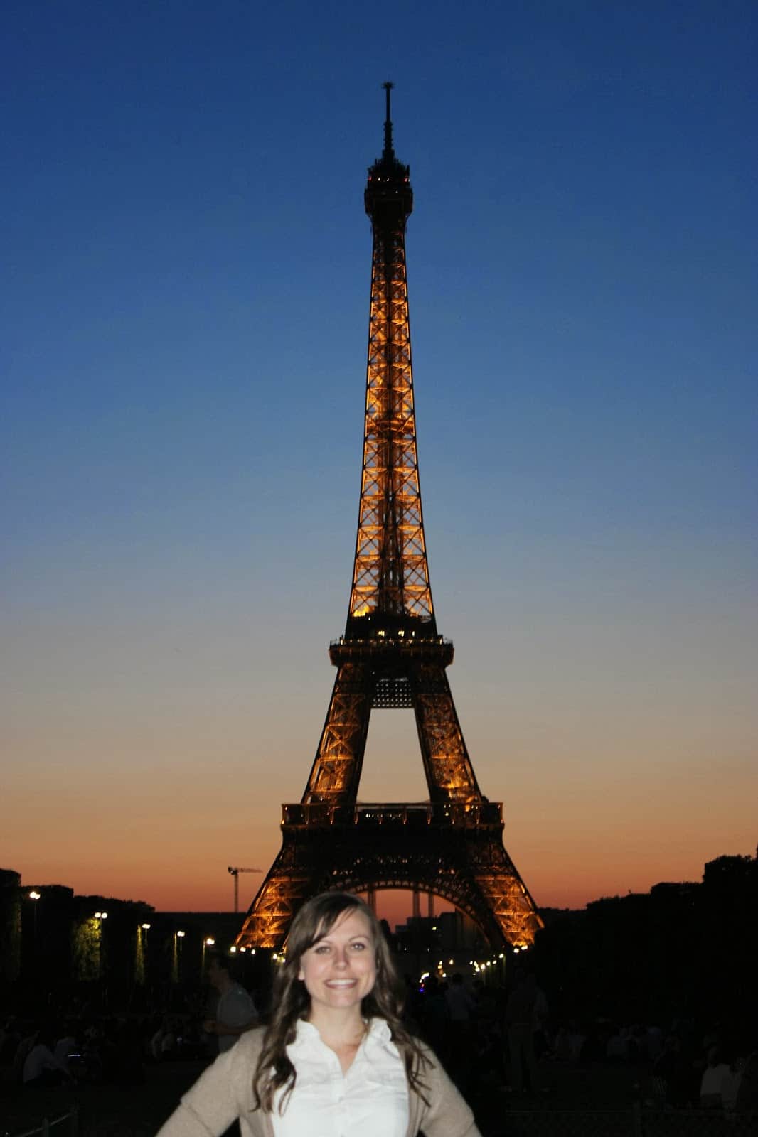 The Eiffel Tower lit up at night with woman smiling in front of it. 