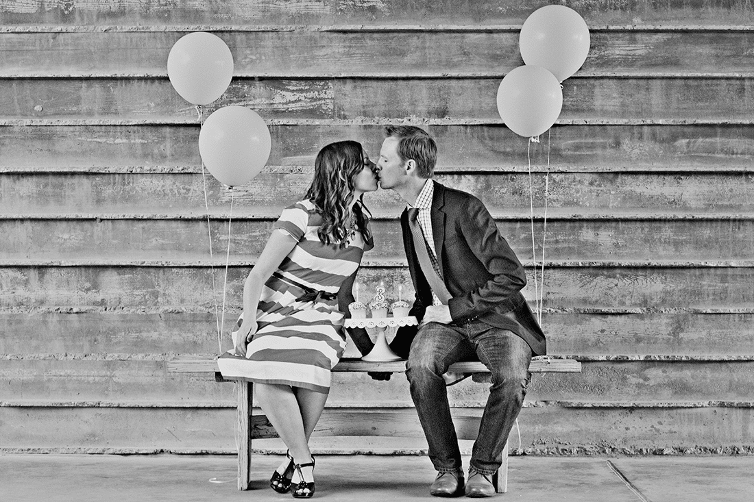 Couple kissing on a bench with balloons behind them. 