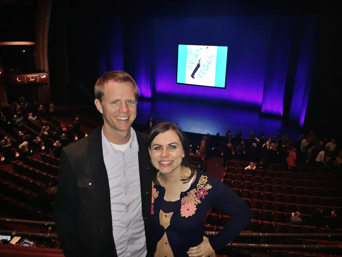 A couple standing in front of the sign at a Brian Regan show. 