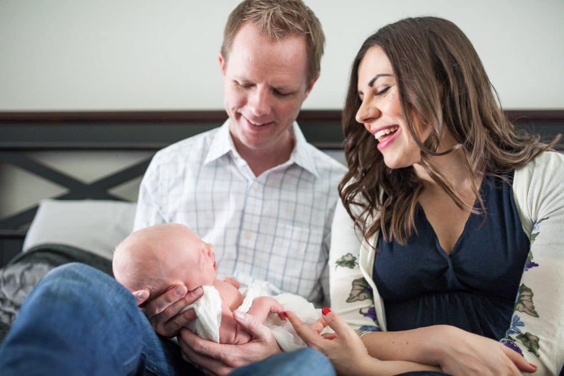 Mom and dad sitting on bed looking at newborn baby. 