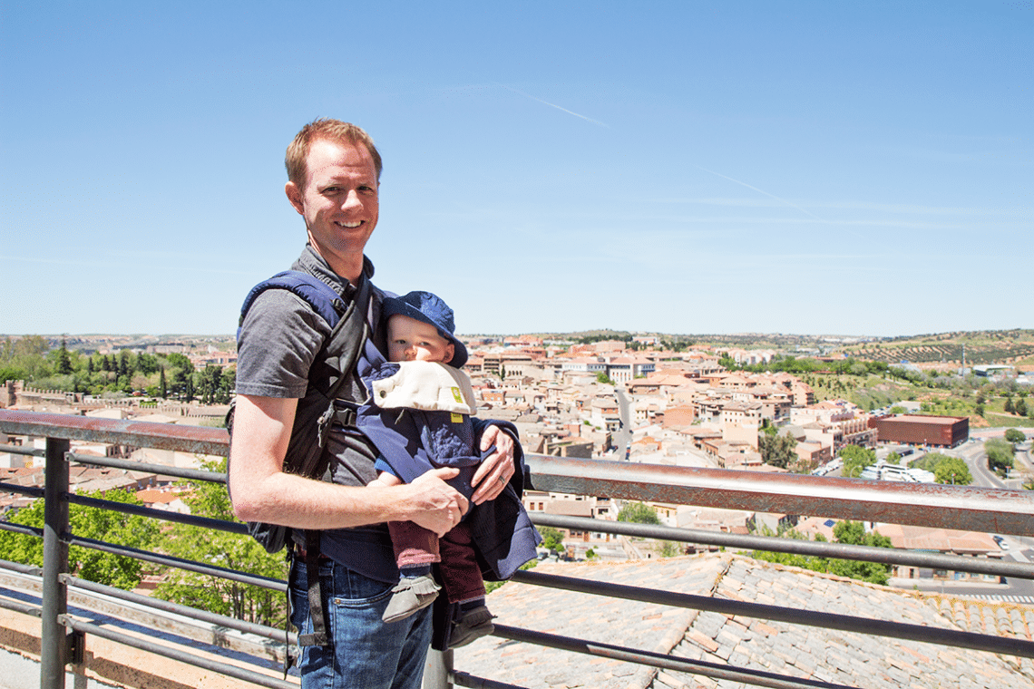 Young dad holding a baby in a baby carrier next to the Toledo Spain skyline. 