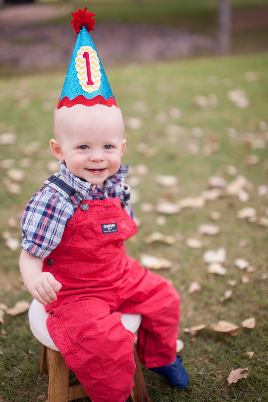First birthday photo shoot: baby in red overalls wearing a birthday hat. 