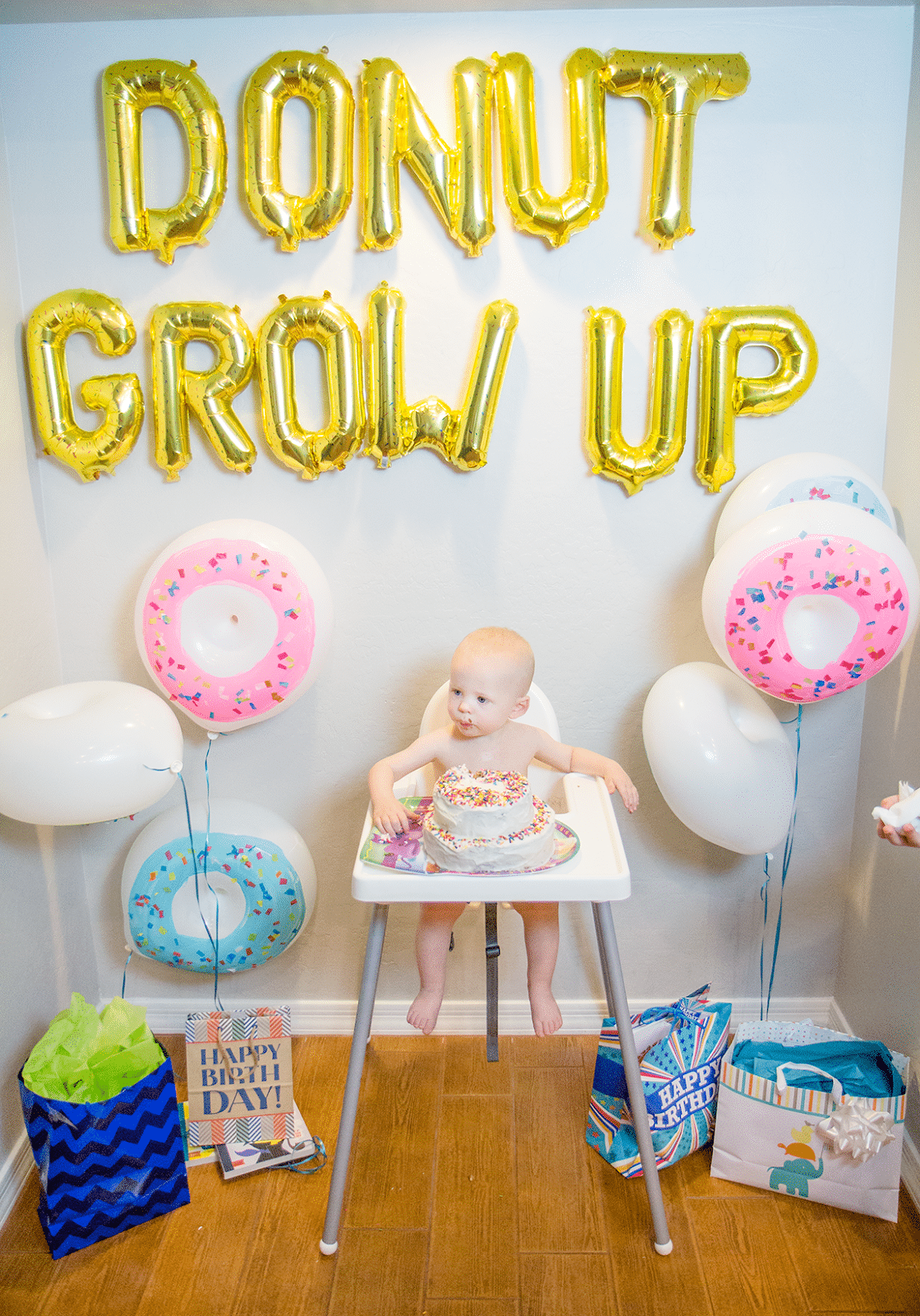 donut grow up gold foil balloons hanging on wall behind a baby in a high chair. 