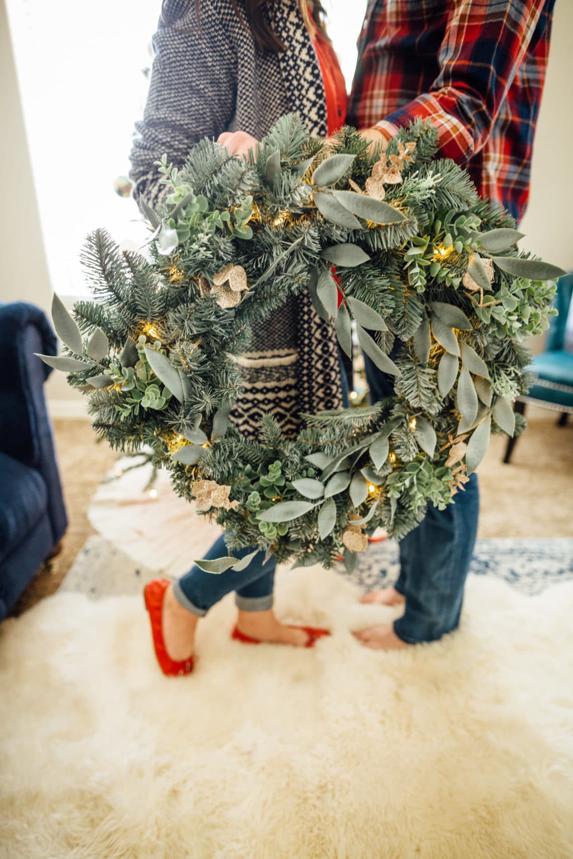 A couple holding up a Christmas wreath leaning into each other to share Christmas Traditions for Couples. 