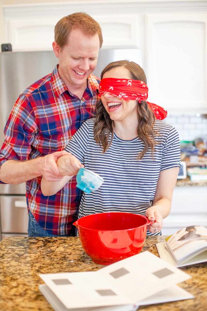Couple baking together for date night. One partner is blindfolded while the other assists them to complete baking tasks. 
