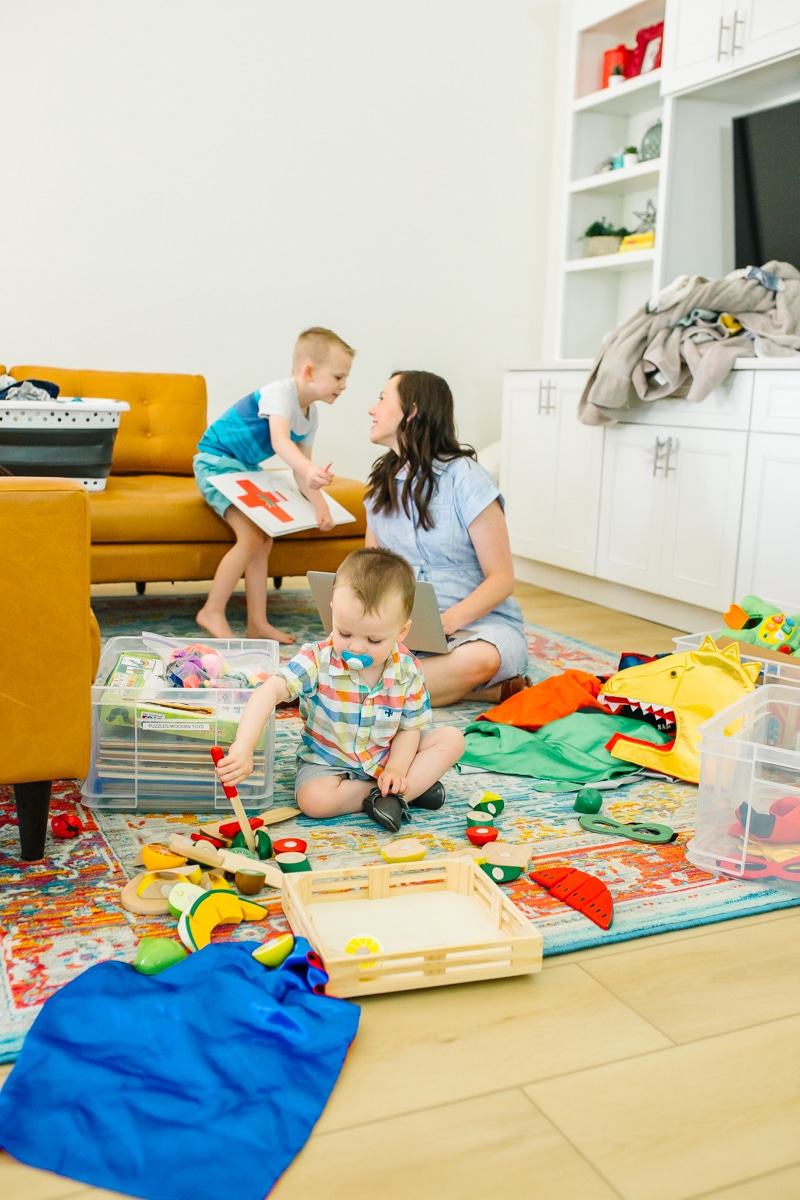 Busy working mom sitting on the floor with her laptop open while kids roam around her with toys and messes. 