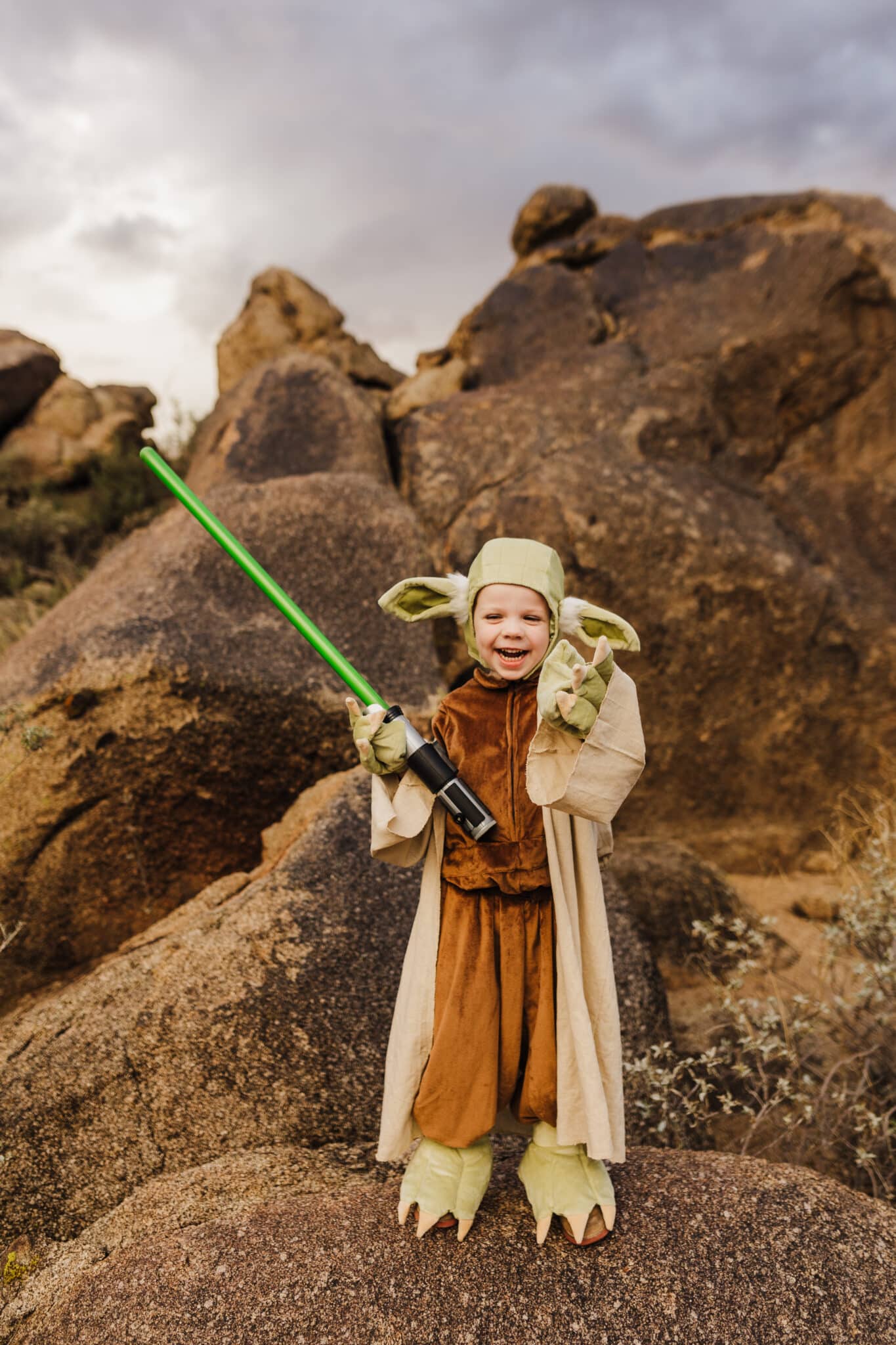 A toddler holding a green lightsaber modeling a Yoda Kids Costume. 