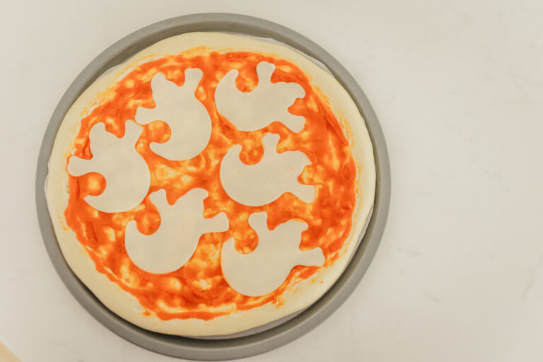 Halloween Pizza with ghost shaped cheese slices on top of pizza dough with sauce.