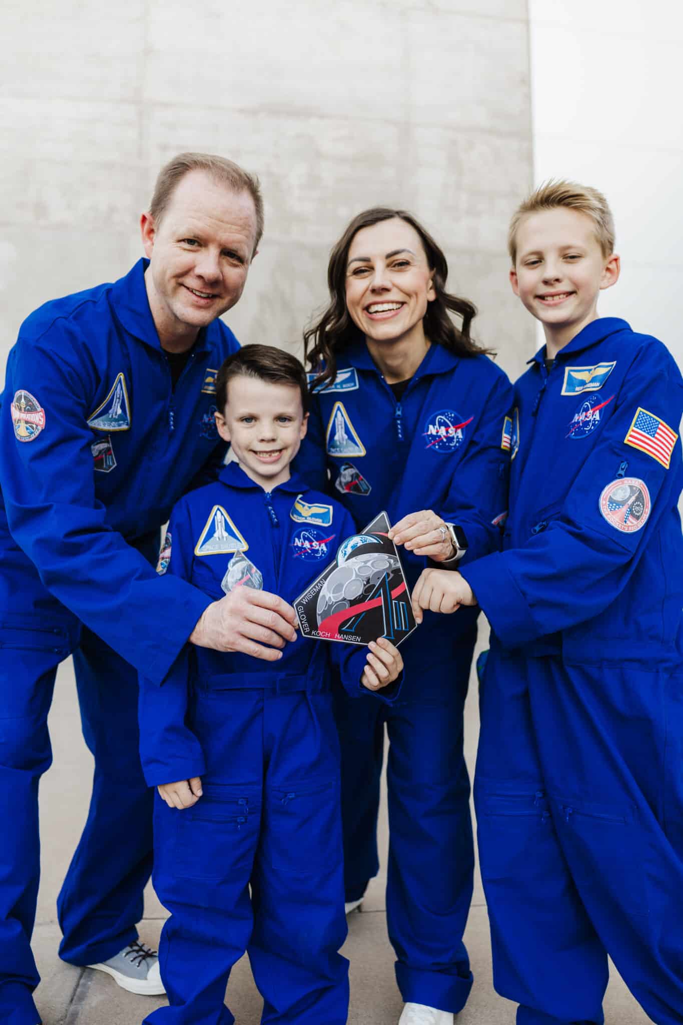 Family astronaut costume: mom, dad, and two boys in blue NASA flight suits holding a giant Artemis II crew patch.