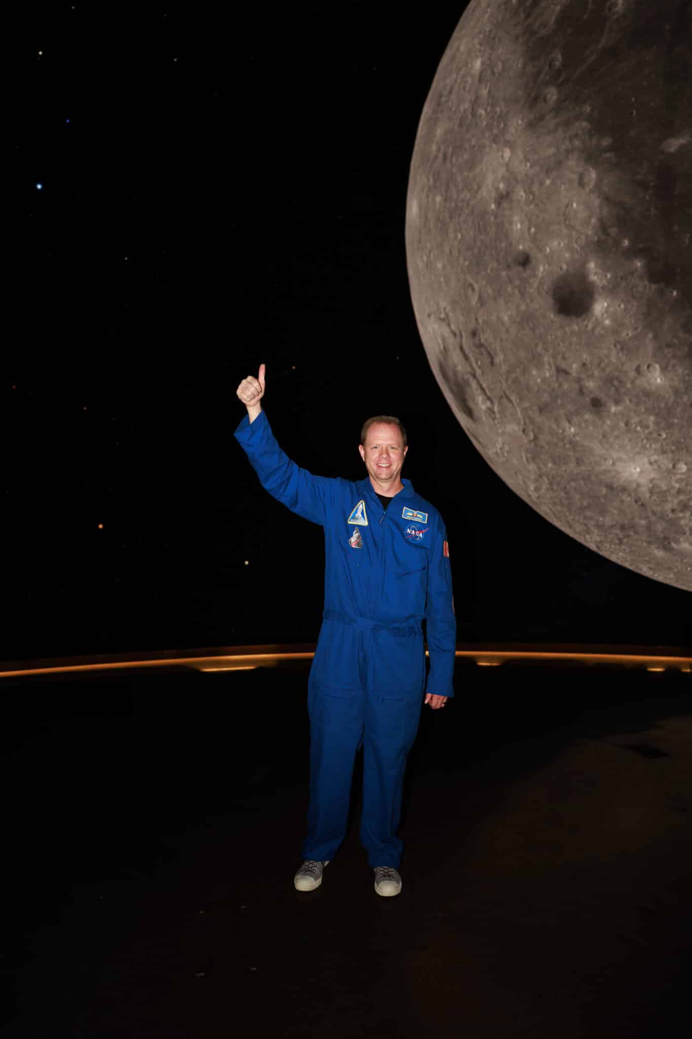 Man in a blue flight suit with a Canadian flag patch holding the Rise plush and giving a thumbs up overhead next to the moon
