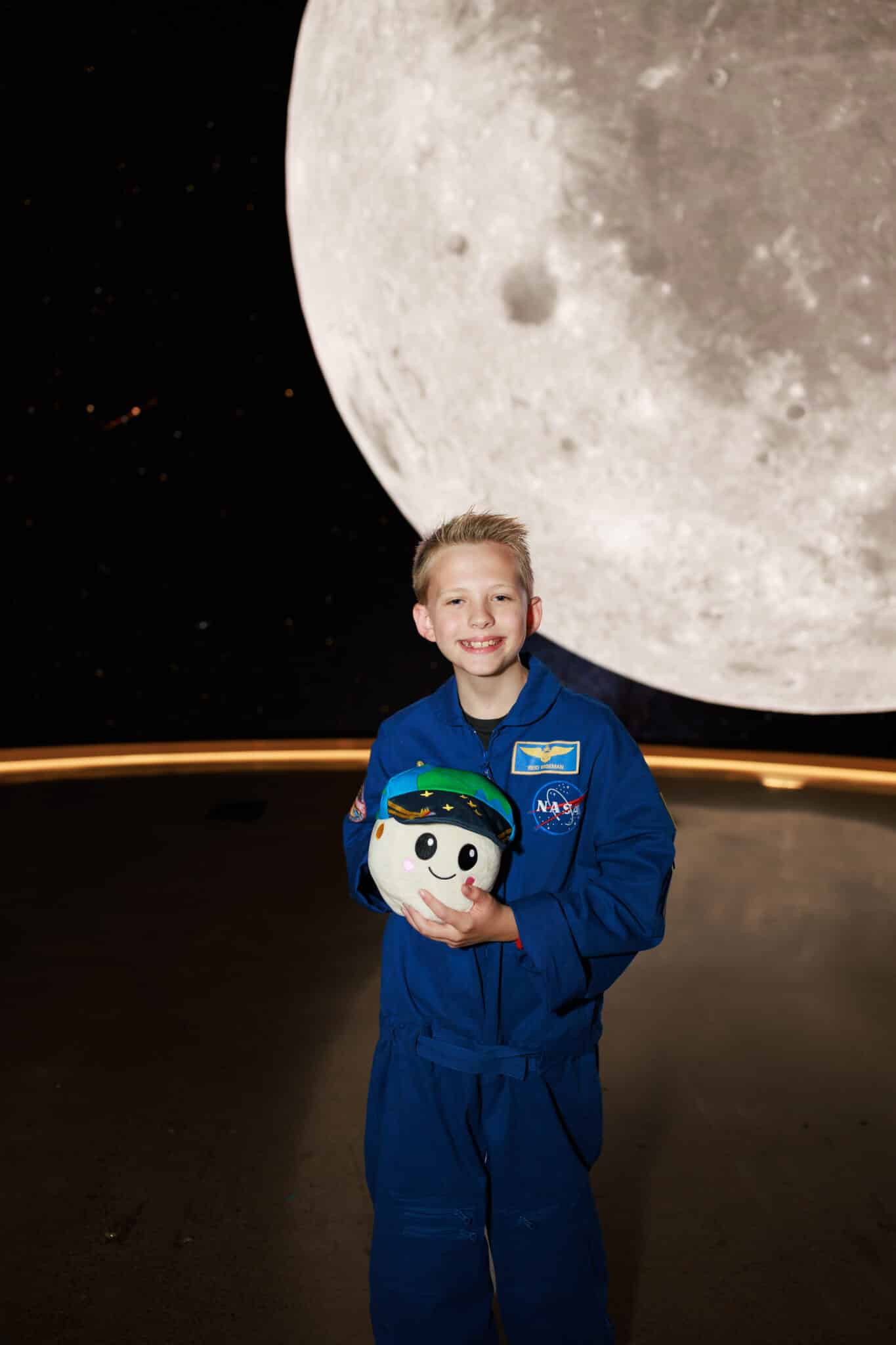 Boy in a blue NASA flight suit as Reid Wiseman holding the Rise plush in front of a giant moon