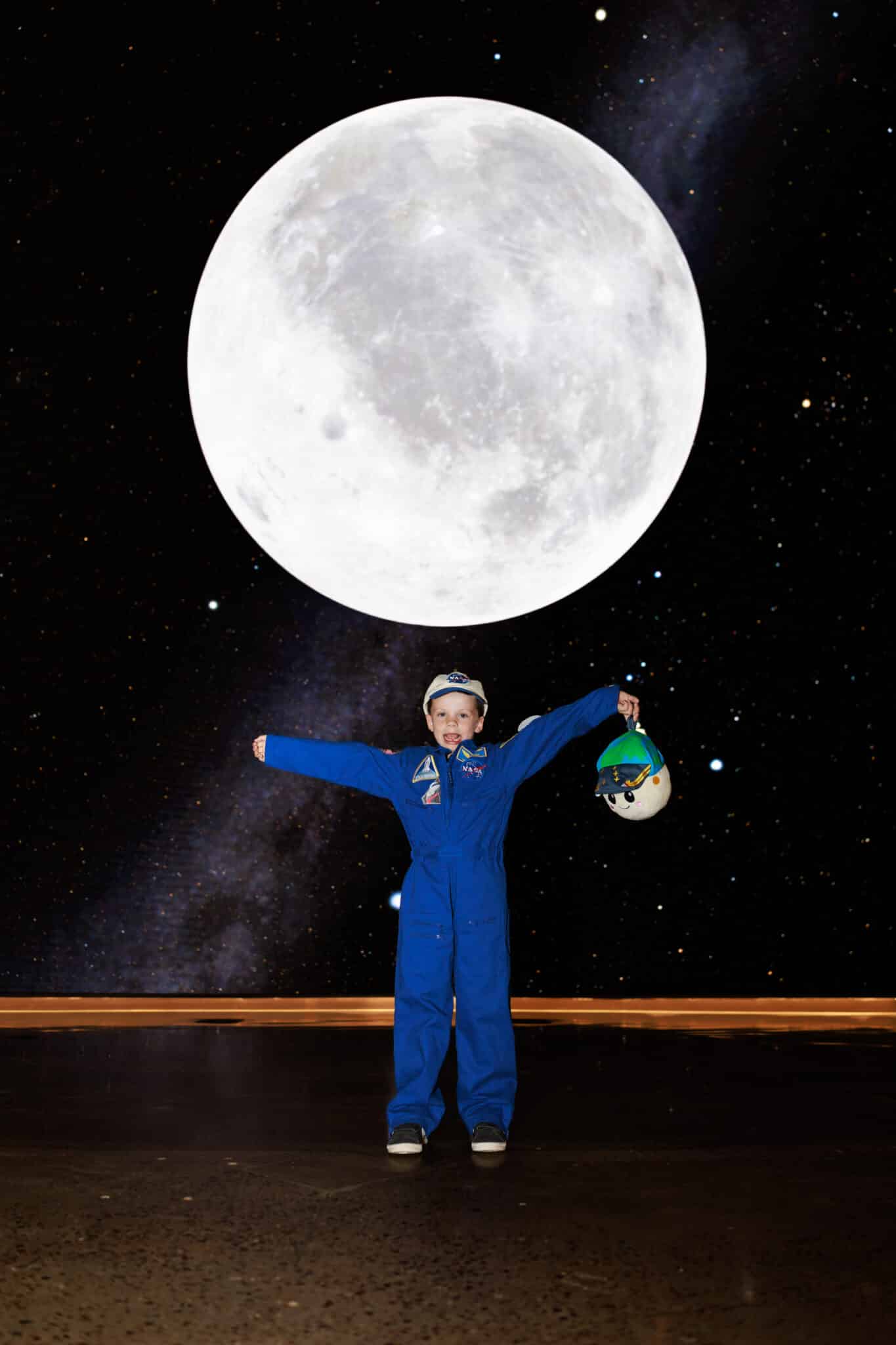 Young boy in a blue flight suit and NASA cap as Victor Glover, arms outstretched under a full moon, holding Rise