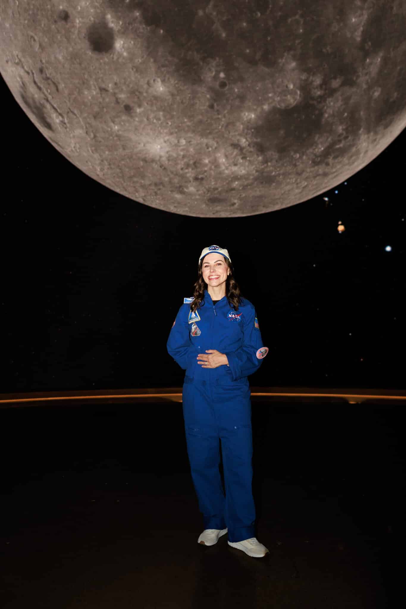 Woman in a blue NASA flight suit and cap as astronaut Christina Koch standing beneath a giant moon