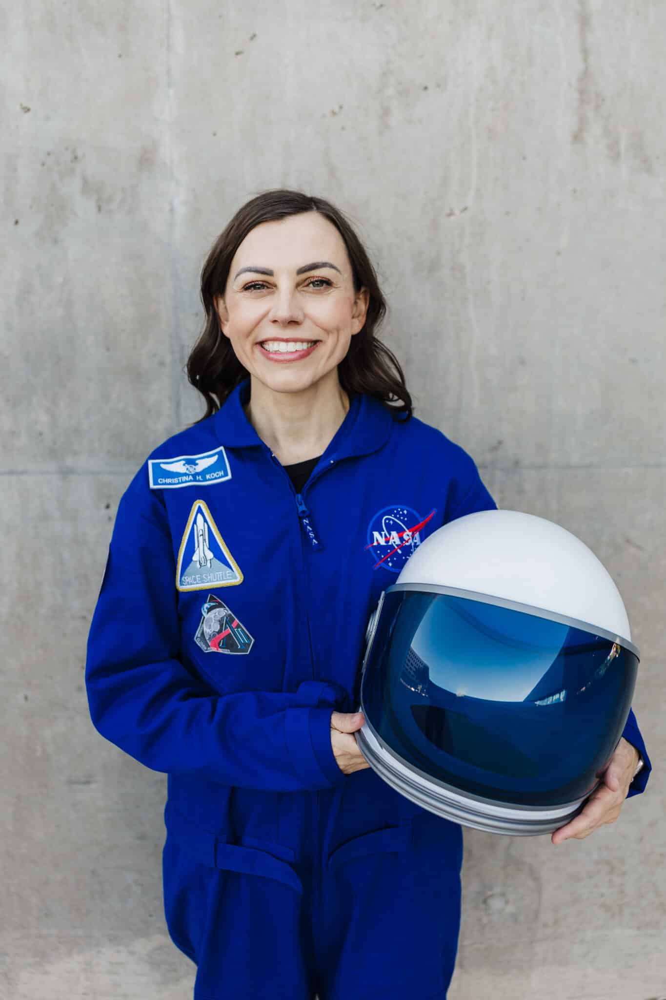 Woman in a blue NASA flight suit holding an astronaut helmet posing as astronaut Christina Koch.