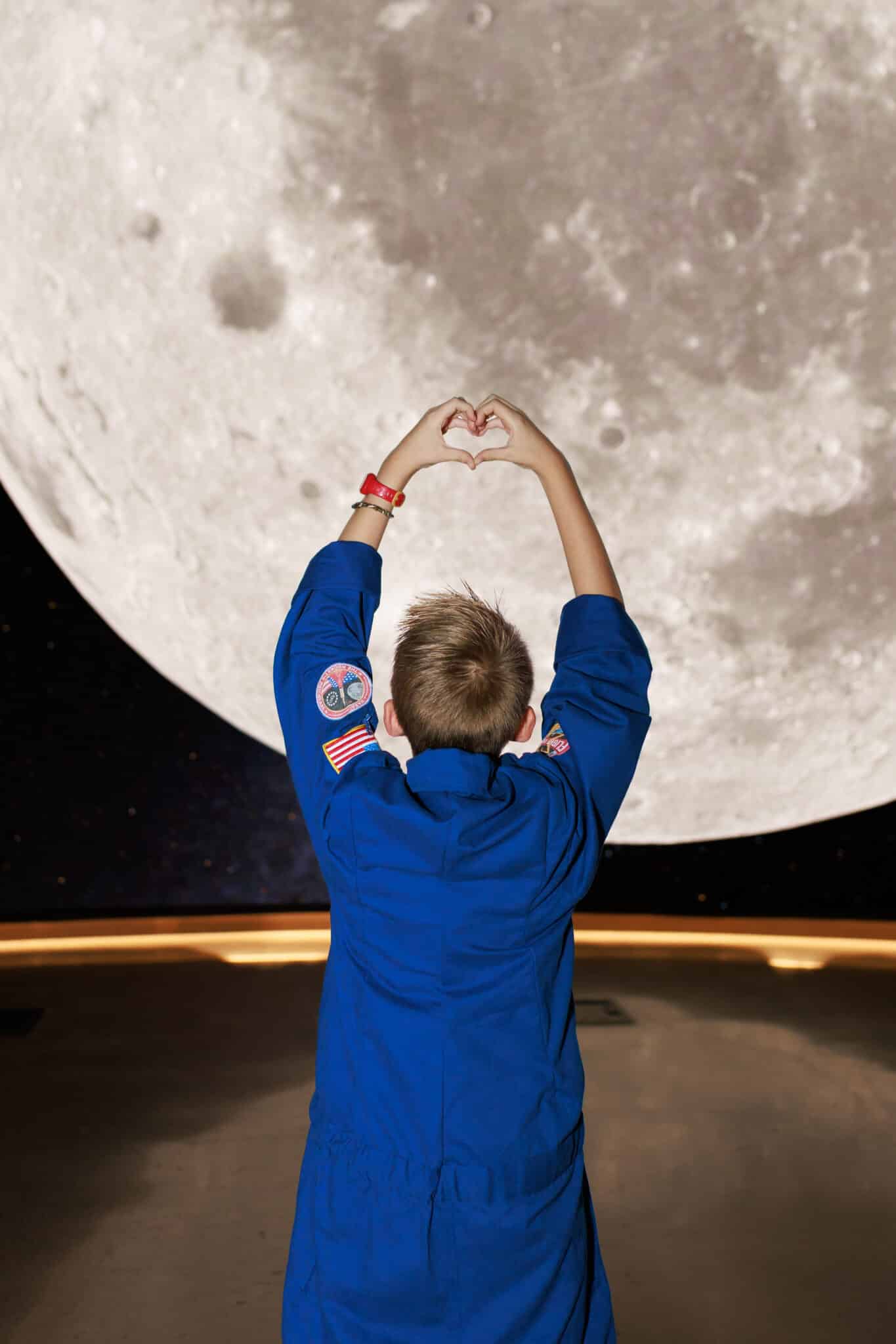 Boy in a blue astronaut flight suit with flag patches dressed as Reid Wiseman making a heart with his hands toward the moon