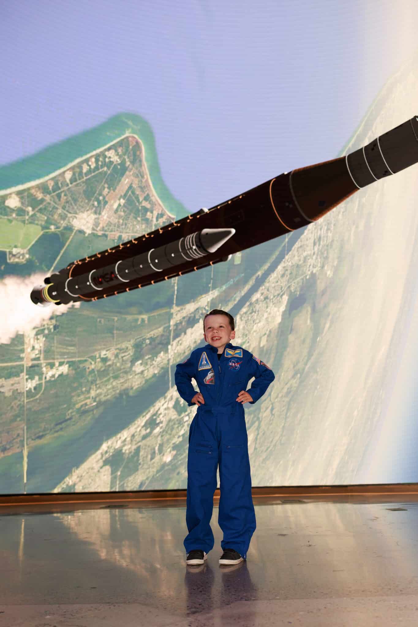 Young boy in a blue flight suit and NASA cap as Victor Glover, arms outstretched under a full moon, holding Rise